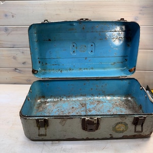 May include: An open, vintage metal tackle box with a light blue interior and a weathered, gray exterior. The box has a hinged lid and metal latches, showing signs of age and use. A circular sticker is visible on the front.