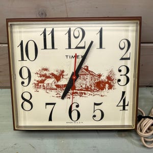 May include: A square wall clock with a brown frame and a cream-colored face featuring large black numerals. The center displays a reddish-brown illustration of a house and trees. It has black hour and minute hands, a red second hand, and the text TIMEX.