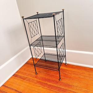 May include: A black metal three-tiered shelf with diamond-patterned side panels. The shelves are made of a mesh material. The top of the shelf has a solid top. The shelf has four small, round, gold-colored feet.
