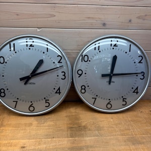 May include: Two round wall clocks with silver frames and light gray faces. Each clock features black Arabic numerals from 1 to 12, black hour and minute hands, and the word STANDARD printed in black below the center. The clocks have glass covers.