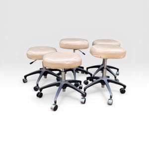 May include: Five cream-colored, adjustable height stools with a rounded seat and a five-point base. The bases are dark gray with rolling wheels. The stools are arranged in a group, with a white background.