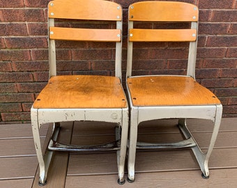 May include: Two vintage school chairs with wooden seats and backs, painted in a light gray color. The chairs have metal frames and are in good condition.