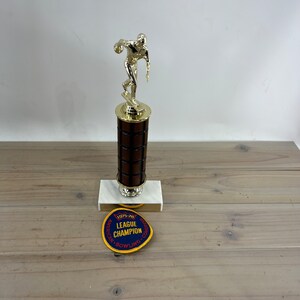 May include: A gold-colored bowling trophy with a wooden base. The trophy features a figure of a bowler in mid-throw. The base has a plaque that reads "1975-76 American Bowling Congress League Champion".