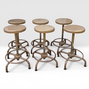 May include: A set of six industrial stools with round, beige-painted metal seats and matching frames. Each stool features a central adjustable post, a circular footrest, and three curved legs with worn paint showing signs of age and use.