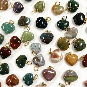 May include: A collection of heart-shaped gemstone pendants in various colors, including green, brown, red, and purple. Each pendant has a gold-colored loop for attaching to a chain or cord. The gemstones have a polished, smooth surface.