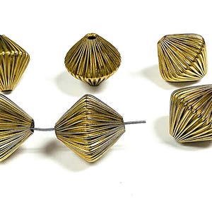 Puede incluir: Primer plano de varias cuentas geométricas doradas. Las cuentas tienen forma de diamante acanalado, con un agujero que atraviesa el centro. Algunas cuentas están ensartadas en un alambre fino. Las cuentas están sobre un fondo blanco.