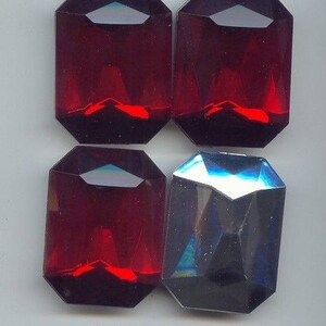 Op de afbeelding: Vier gefacetteerde glazen edelstenen, drie zijn rood en één is blauw. De edelstenen zijn gevormd als langwerpige achthoeken.