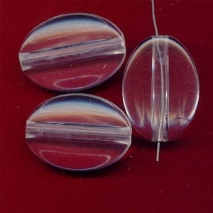 May include: Three clear, oval-shaped beads with a horizontal groove. The beads have a subtle iridescent sheen, reflecting shades of white, blue, and orange. One bead is strung on a thin, white thread. The beads are on a red background.