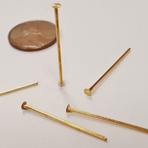 May include: Five gold-colored head pins with flat heads. The pins are different lengths and are arranged on a white surface. A penny is in the background for size comparison.