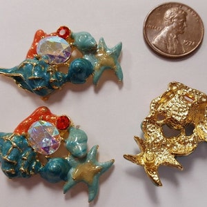 Puede incluir: Dos abalorios de metal dorado en tono dorado con temática marina con esmalte azul, verde y naranja. Cada abalorio presenta una estrella de mar, conchas marinas y una pedrería transparente iridiscente.
