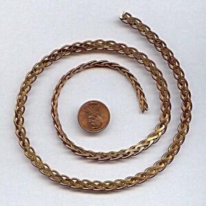 Op de afbeelding: Een gouden ketting met een gevlochten ontwerp. De ketting is opgerold en gelegd op een wit oppervlak. Een cent is naast de ketting gelegd ter vergelijking van de grootte.