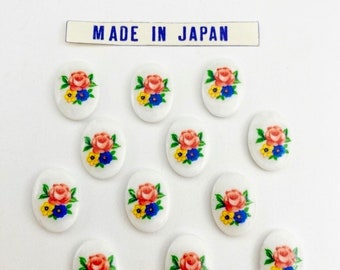 May include: Twelve oval-shaped white buttons with a floral design. Each button features a pink rose with yellow and blue flowers. The buttons are labeled "MADE IN JAPAN".