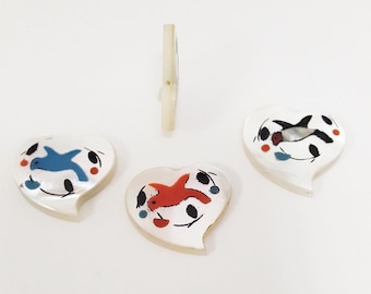 May include: Three heart-shaped buttons with white backgrounds and colorful painted fish designs. The fish are blue, red, and black.
