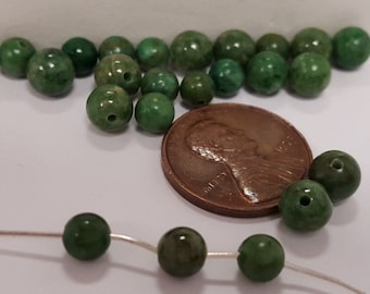 Op de afbeelding: Een verzameling groene edelsteen kralen, sommige geregen aan een zilveren draad, naast een Amerikaanse penny ter groottevergelijking.