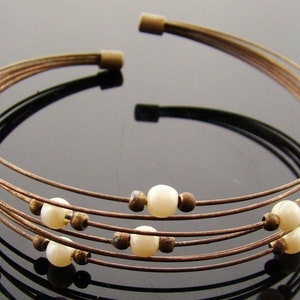 May include: A multi-strand bracelet made of brown wire with white pearl beads. The bracelet is open and has a simple design.