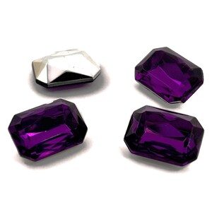 Puede incluir: Cuatro gemas de vidrio púrpura con un diseño facetado. Las gemas son de forma rectangular y tienen un acabado brillante.