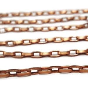 May include: A close-up of a copper chain with a flat, oval link design. The chain is lying on a white surface.