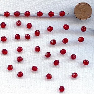 May include: A collection of faceted, deep red beads scattered on a white surface. Some beads are strung on a thin wire. A US penny is included for size reference. The beads have a sparkling, gem-like appearance, suitable for jewelry making.