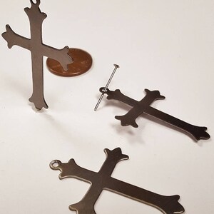 May include: Three silver metal cross charms with ornate details. The crosses are different sizes and have a hole at the top for hanging.