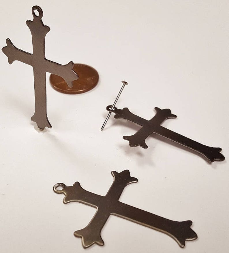 May include: Three silver metal cross charms with ornate details. The crosses are different sizes and have a hole at the top for hanging.