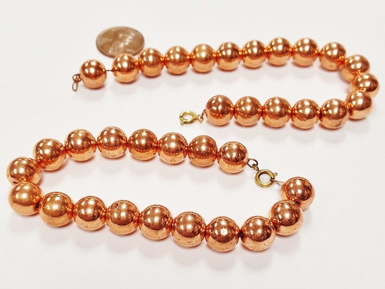 May include: A copper bead necklace with a shiny, metallic finish. The necklace is made of round beads and has a simple design.