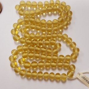 Puede incluir: Una hebra de cuentas de vidrio amarillo con un aspecto claro y ligeramente nublado. Las cuentas son redondas y tienen una ligera textura. La hebra est&aacute; etiquetada con "IN JAPAN".