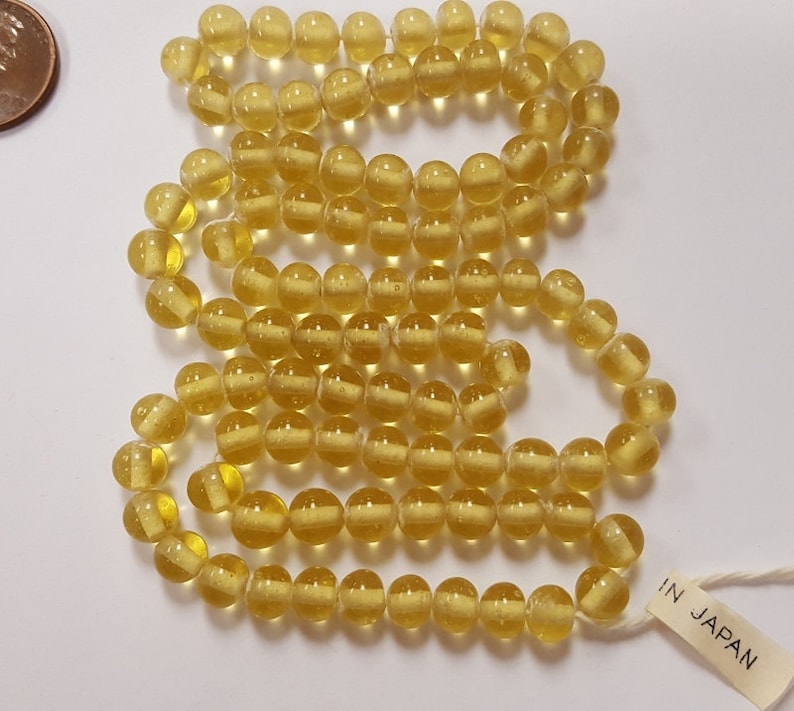 Puede incluir: Una hebra de cuentas de vidrio amarillo con un aspecto claro y ligeramente nublado. Las cuentas son redondas y tienen una ligera textura. La hebra est&aacute; etiquetada con "IN JAPAN".