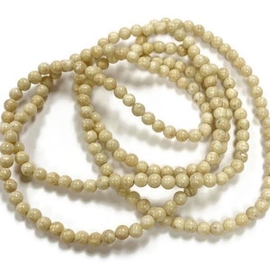 May include: A strand of cream-colored round beads, made of fossilized coral, is shown in a close-up image. The beads are arranged in a circular pattern, with several strands overlapping.