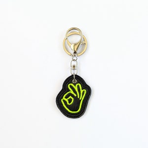 May include: A black leather keychain with a neon green outline of an OK hand gesture.