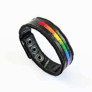 Könnte beinhalten: Schwarzes Lederarmband mit einem Regenbogenstreifen-Design. Das Armband hat drei Metallösen.