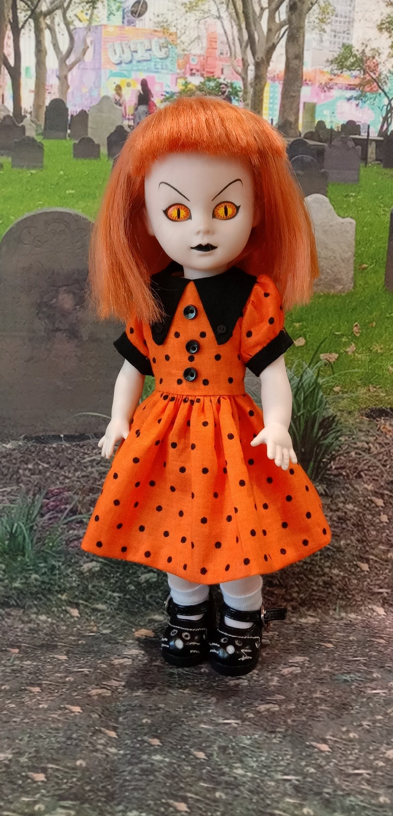 May include: A doll with orange hair and yellow eyes, wearing an orange dress with black polka dots and black shoes. The doll has a black collar and is posed in front of a cemetery.