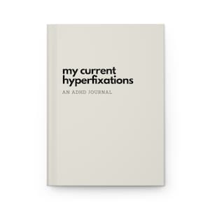 Op de afbeelding: Een beige notitieboek met de tekst "my current hyperfixations" in zwart, en de woorden "AN ADHD JOURNAL" in kleinere zwarte tekst eronder.