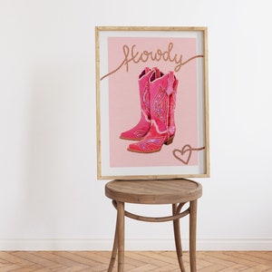 K&ouml;nnte beinhalten: Ein gerahmter Druck mit rosa Hintergrund, der ein Paar rosa Cowboystiefel mit glitzernden Verzierungen zeigt. Der Text "Howdy" ist in brauner Kursivschrift &uuml;ber den Stiefeln geschrieben. Ein kleines rosa Herz ist in der rechten unteren Ecke gezeichnet.