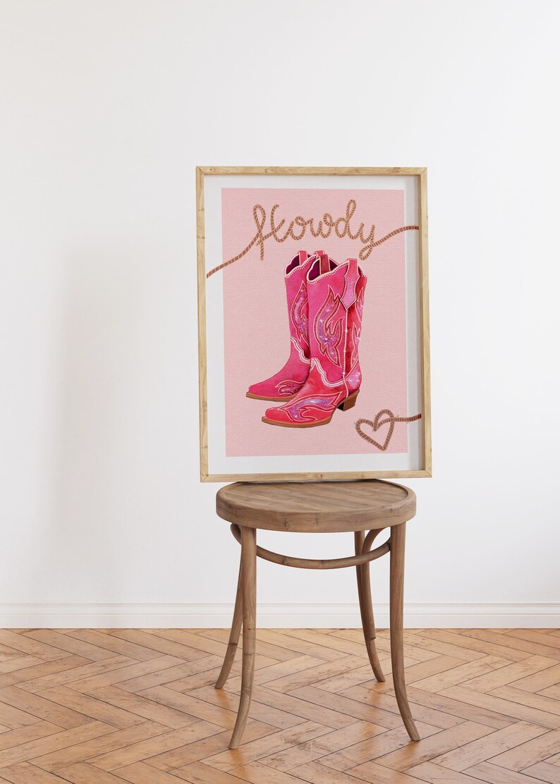 K&ouml;nnte beinhalten: Ein gerahmter Druck mit rosa Hintergrund, der ein Paar rosa Cowboystiefel mit glitzernden Verzierungen zeigt. Der Text "Howdy" ist in brauner Kursivschrift &uuml;ber den Stiefeln geschrieben. Ein kleines rosa Herz ist in der rechten unteren Ecke gezeichnet.