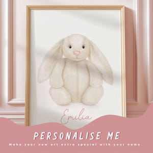 Puede incluir: Una lámina de arte enmarcada con la ilustración de un conejo de peluche color crema sobre fondo blanco. Debajo del conejo, el nombre Emilia aparece escrito en letra cursiva de color rosa. El marco es de un tono madera claro.