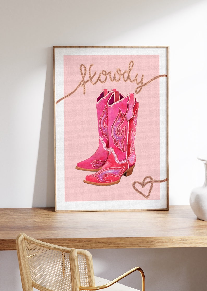 K&ouml;nnte beinhalten: Ein rosa Hintergrund mit dem Wort "Howdy" in braunem Seil geschrieben. Zwei rosa Cowboystiefel mit glitzernden Details befinden sich auf dem Hintergrund. Ein braunes Seilherz befindet sich in der rechten unteren Ecke.