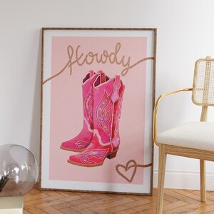 K&ouml;nnte beinhalten: Ein rosa Poster mit dem Wort "Howdy" in braunem Seil geschrieben. Das Poster zeigt ein Paar rosa Cowboystiefel mit glitzernden Details. Ein braunes Seilherz befindet sich am unteren Rand des Posters.
