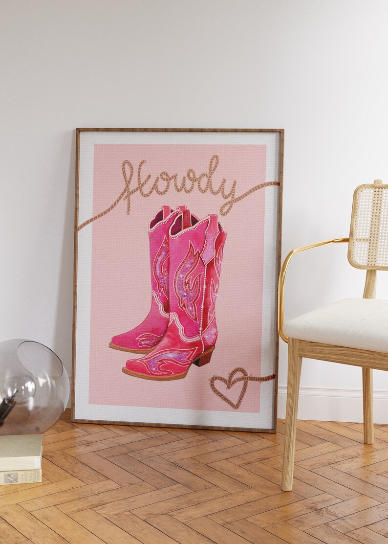 K&ouml;nnte beinhalten: Ein rosa Poster mit dem Wort "Howdy" in braunem Seil geschrieben. Das Poster zeigt ein Paar rosa Cowboystiefel mit glitzernden Details. Ein braunes Seilherz befindet sich am unteren Rand des Posters.
