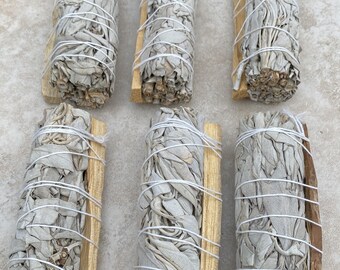 May include: Six bundles of dried white sage leaves arranged in two rows. Each bundle is tightly wrapped with thin white string and secured alongside a single, light brown wooden stick. The sage leaves are pale gray-green and textured.