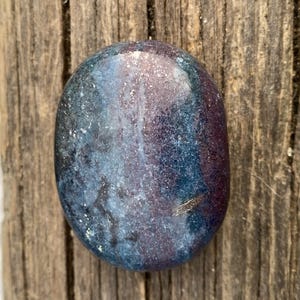 May include: An oval-shaped, polished stone with a blend of blue, purple, and white hues. The stone has a smooth, glossy surface and is set against a backdrop of weathered wood. The colors create a marbled effect.