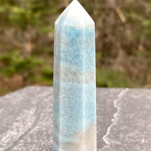 May include: A tall, hexagonal crystal point with a pointed top. The crystal features bands of light blue, white, and tan colors. The crystal is standing on a gray surface, with a blurred green background.