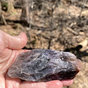 K&ouml;nnte beinhalten: Ein roher, nat&uuml;rlicher Amethystkristall mit lila, grauen und braunen Farbt&ouml;nen. Der Kristall wird in einer Hand gehalten, wodurch seine unregelm&auml;&szlig;ige Form und strukturierte Oberfl&auml;che sichtbar werden. Der Hintergrund ist unscharf, was auf eine Umgebung im Freien hindeutet.