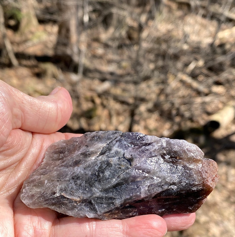 K&ouml;nnte beinhalten: Ein roher, nat&uuml;rlicher Amethystkristall mit lila, grauen und braunen Farbt&ouml;nen. Der Kristall wird in einer Hand gehalten, wodurch seine unregelm&auml;&szlig;ige Form und strukturierte Oberfl&auml;che sichtbar werden. Der Hintergrund ist unscharf, was auf eine Umgebung im Freien hindeutet.