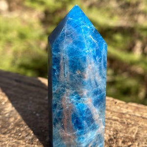 May include: A vibrant blue crystal point with a polished, faceted surface. The crystal has a rectangular base and tapers to a pointed apex. Internal veining adds depth and visual interest to the translucent stone.