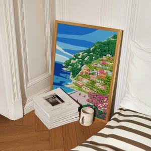 Beach art print with colorful flowers and bright houses and pink and green flowers on a mountainside hill with bright blue sky and little people on the shore and swimming in the bluey green water ocean sea. Calming and whimsical image of visitors to Positano village in Italy Europe. Natural wooden frame of Positano art print original artwork in European style architecture interior home wooden floors and modern bed with magazines and cup of coffee