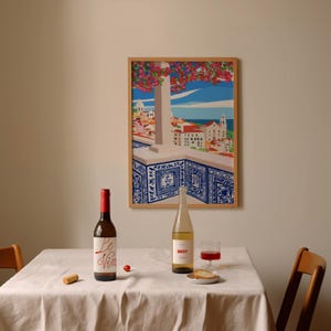 May include: A white tablecloth with two bottles of wine, one red and one white, a glass of red wine, a small plate with bread, and a cork on the table. A framed print of a cityscape with a blue and white tiled balcony hangs on the wall behind the table.