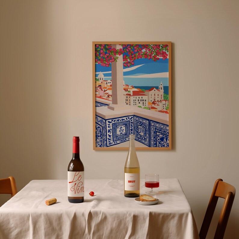May include: A white tablecloth with two bottles of wine, one red and one white, a glass of red wine, a small plate with bread, and a cork on the table. A framed print of a cityscape with a blue and white tiled balcony hangs on the wall behind the table.