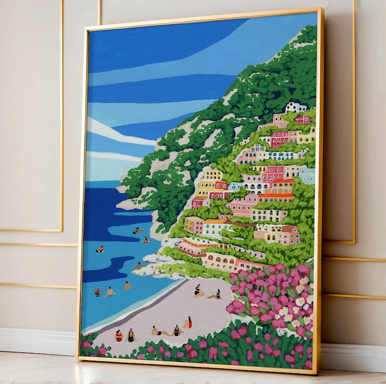 Beach art print with colorful flowers and bright houses and pink and green flowers on a mountainside hill with bright blue sky and little people on the shore and swimming in the bluey green water ocean sea. Calming and whimsical image of visitors to Positano village in Italy Europe. Gold Framed image of Positano leaning up against a Pink and Gold wall in European architecture interior building or home