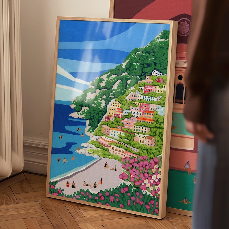 Pu&ograve; includere: Un dipinto colorato di una citt&agrave; costiera con edifici su una collina, una spiaggia e acqua blu. Il dipinto &egrave; incorniciato in una cornice di legno chiaro.