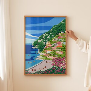 Beach art print with colorful flowers and bright houses and pink and green flowers on a mountainside hill with bright blue sky and little people on the shore and swimming in the bluey green water ocean sea. Calming and whimsical image of visitors to Positano village in Italy Europe. Natural wooden framed art print of positano with human hand and white shirted person touching the frame. Soft and neutral background and curtain
